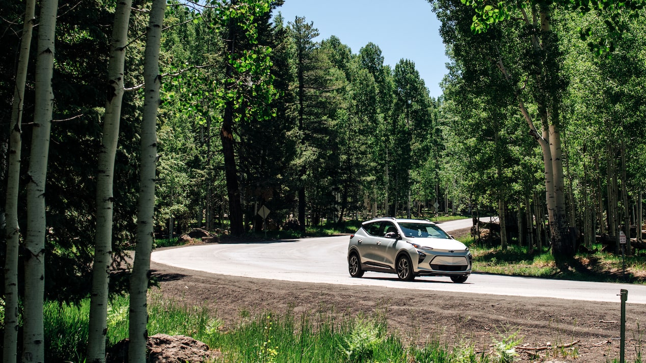 A Chevrolet Bolt EUV drives along a winding road between tall columns of evergreen trees and a blue sky above.