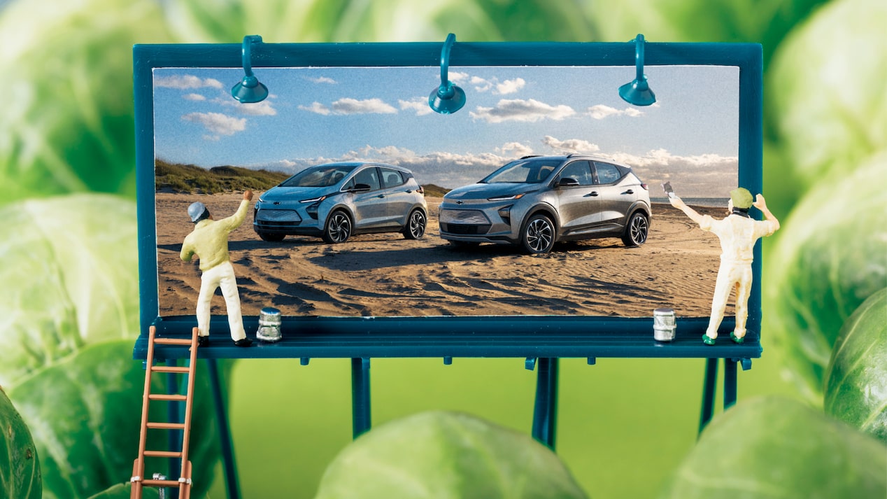 A diorama-like image of a miniature billboard showing the Bolt EV and Bolt EUV, with two tiny human figures painting it, surrounded by Brussels sprouts to emulate trees.