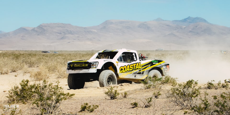 Front Three-Quarters View of Jake Velasco's Off-Roading Truck Driving in the Desert With Mountains in the Background